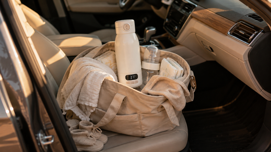Chauffe-biberon nomade LAYNOA dépassant d'un sac à langer beige posé sur le siège passager d'une voiture, avec biberon, lange et chaussons bébé, lumière dorée